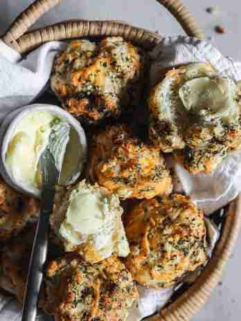 A basket filled with warm cheese biscuits, some halved and topped with butter, alongside a small dish of spreadable butter and a butter knife.