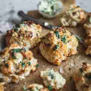 Close-up of golden, flaky biscuits garnished with fresh herbs. One biscuit is broken open, showing its soft interior next to a knife and herb butter.