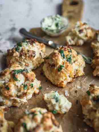 Close-up of golden, flaky biscuits garnished with fresh herbs. One biscuit is broken open, showing its soft interior next to a knife and herb butter.