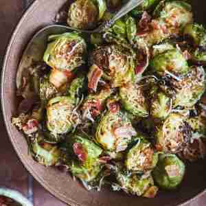 A close-up of roasted Brussels sprouts mixed with crispy pancetta and topped with melted Romano cheese, served in a bowl with a spoon for easy sharing.