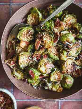 A close-up of roasted Brussels sprouts mixed with crispy pancetta and topped with melted Romano cheese, served in a bowl with a spoon for easy sharing.