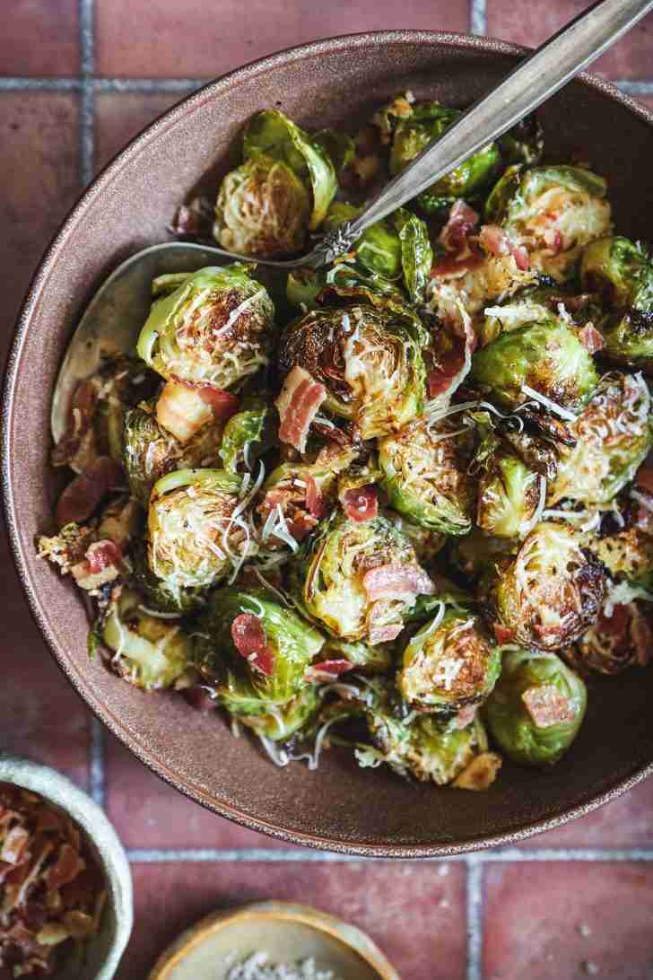 A close-up of roasted Brussels sprouts mixed with crispy pancetta and topped with melted Romano cheese, served in a bowl with a spoon for easy sharing.