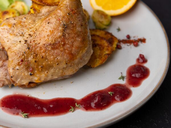 Duck confit served with roasted potatoes, Brussels sprouts, a fresh orange slice, and a drizzle of berry sauce