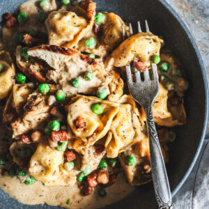 Close-up of chicken tortellini in creamy Alfredo sauce with peas and pancetta, served in a bowl with a fork.