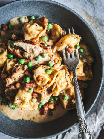 Close-up of chicken tortellini in creamy Alfredo sauce with peas and pancetta, served in a bowl with a fork.