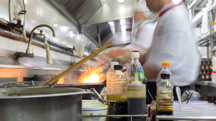 Busy restaurant kitchen with chefs cooking, flames from a wok, and bottles of oils and sauces in the foreground