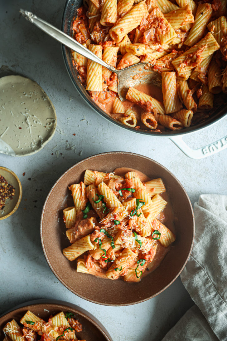 Bowls of rigatoni in creamy vodka sauce topped with fresh basil and Parmesan, served alongside a skillet of pasta and chili flakes.