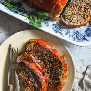 wo slices of glazed meatloaf on a plate with a fork and knife, next to the remaining loaf on a floral platter garnished with fresh parsley.