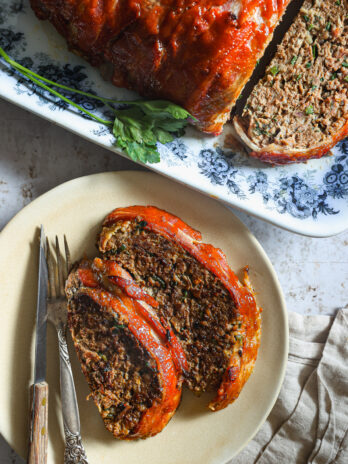 wo slices of glazed meatloaf on a plate with a fork and knife, next to the remaining loaf on a floral platter garnished with fresh parsley.