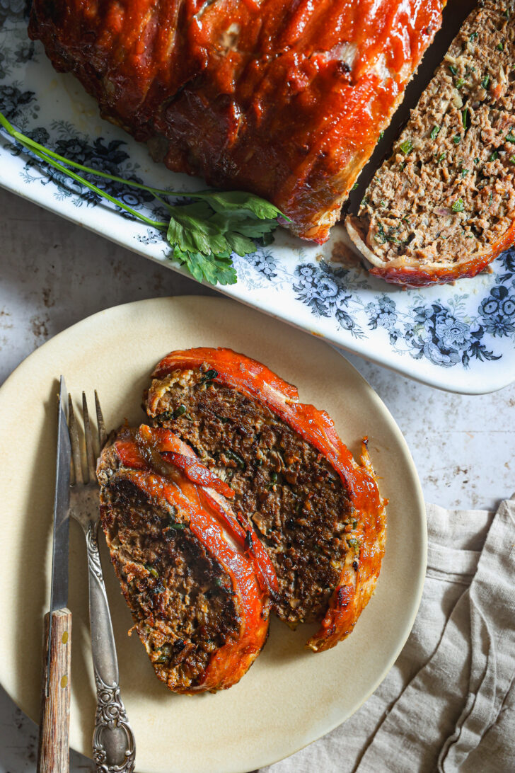 wo slices of glazed meatloaf on a plate with a fork and knife, next to the remaining loaf on a floral platter garnished with fresh parsley.