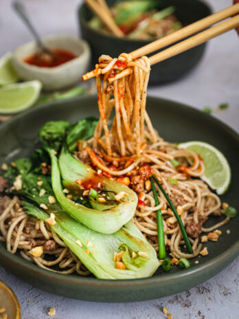 Chopsticks lifting spicy noodles from a bowl of Dan Dan noodles, garnished with bok choy, peanuts, scallions, and lime