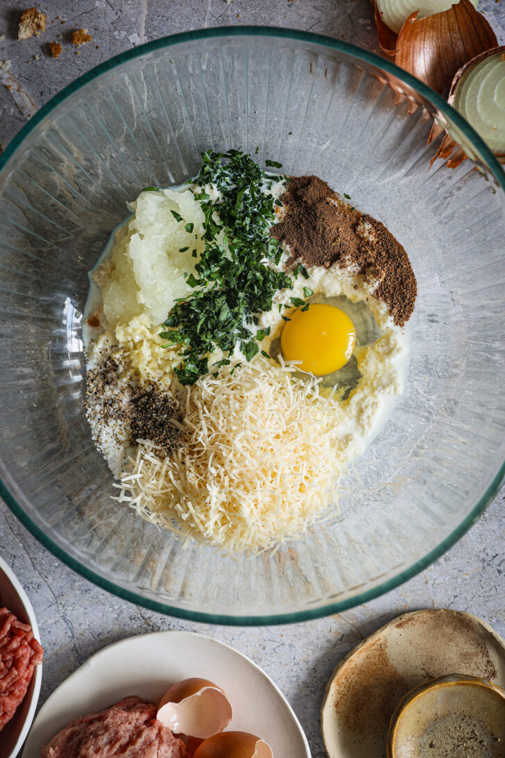 Ingredients for meatball mixture in a glass bowl, including egg, parsley, grated onion, Parmesan cheese, breadcrumbs, and spices.