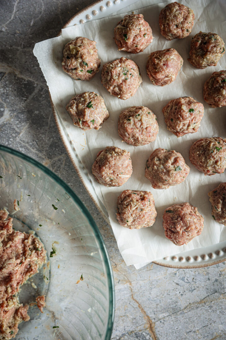 Shaped meatballs arranged on parchment paper, ready for cooking, with the remaining meat mixture in a glass bowl nearby.