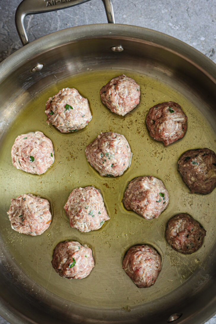 Meatballs browning in a skillet with olive oil, showing a golden crust forming on some as they cook.