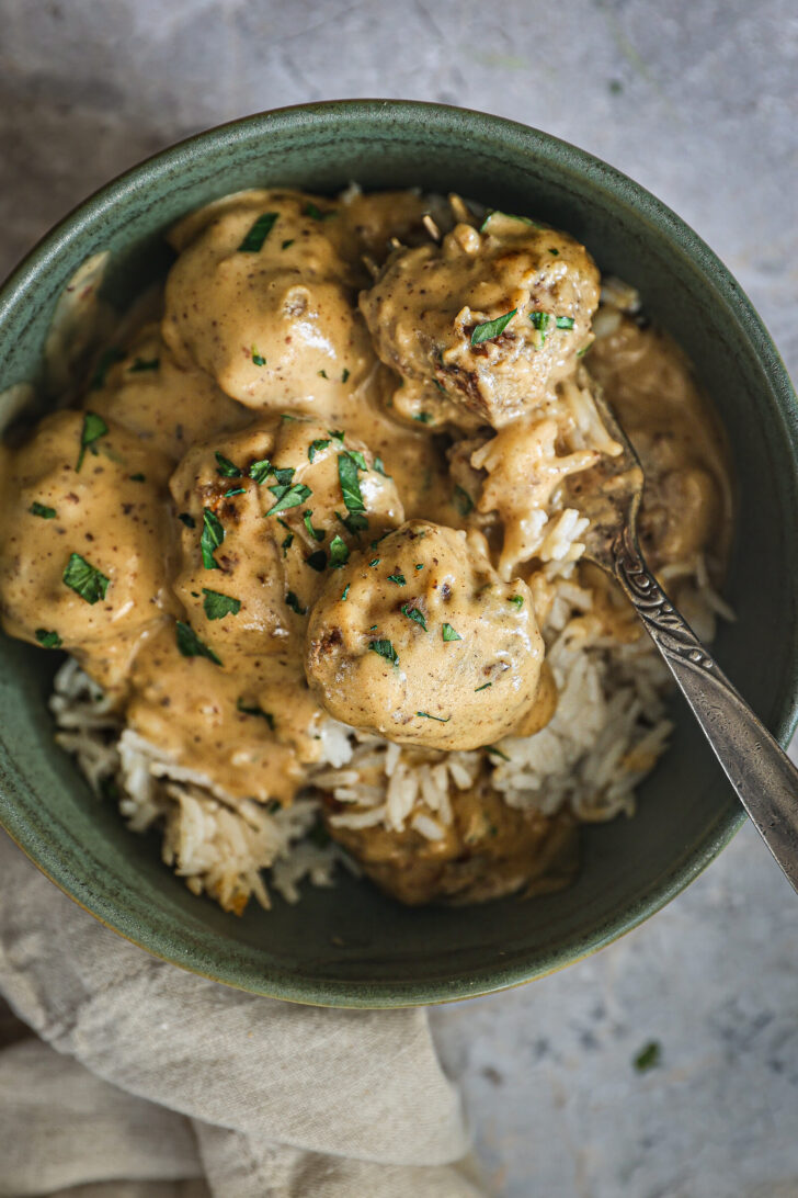 A bowl of Swedish meatballs served over white rice, topped with creamy gravy and fresh parsley, with a fork ready for eating.