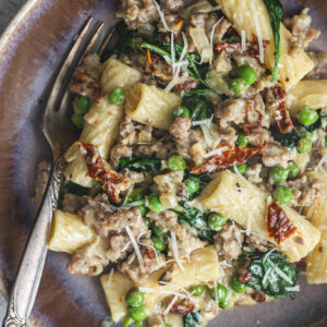 A bowl of creamy Tuscan Rigatoni, loaded with sausage, spinach, peas, and sun-dried tomatoes, topped with shredded Parmesan cheese