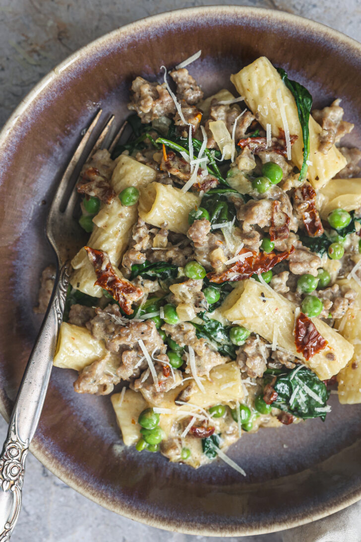 A bowl of creamy Tuscan Rigatoni, loaded with sausage, spinach, peas, and sun-dried tomatoes, topped with shredded Parmesan cheese