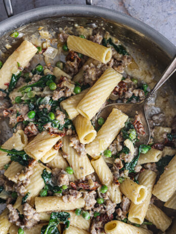 Creamy Tuscan Rigatoni in a skillet, featuring tender pasta, sausage, spinach, peas, and sun-dried tomatoes, ready to serve