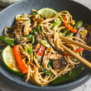 Chopsticks lift beef lo mein with shiitake mushrooms, snap peas, and red bell peppers, garnished with sesame seeds, scallions, and lime