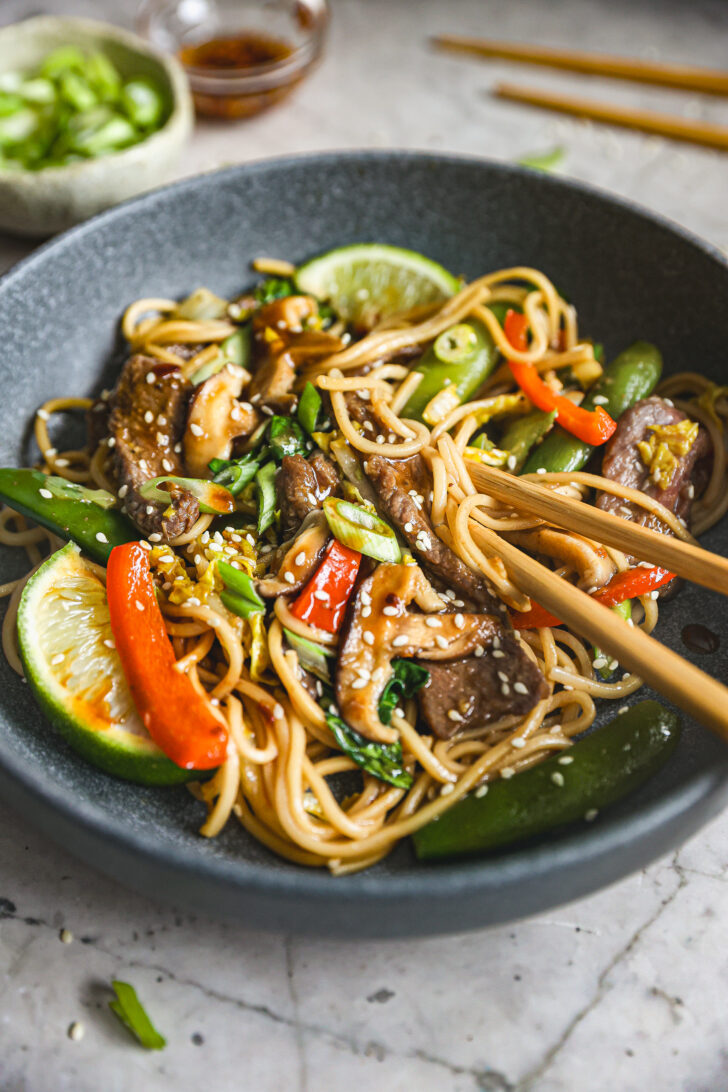 Chopsticks lift beef lo mein with shiitake mushrooms, snap peas, and red bell peppers, garnished with sesame seeds, scallions, and lime