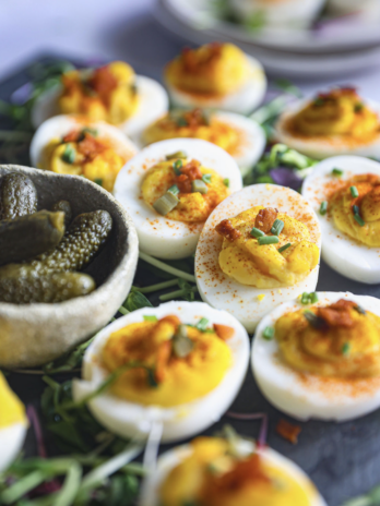 Deviled eggs topped with paprika, chives, bacon, and pickles, served on microgreens with a rustic bowl of gherkins in the foreground