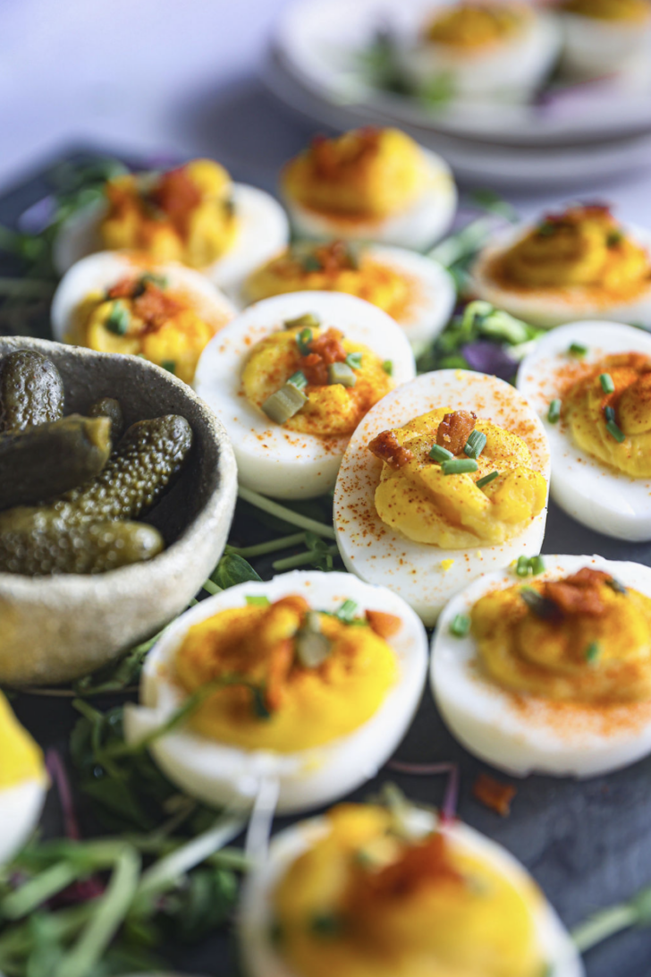 Deviled eggs topped with paprika, chives, bacon, and pickles, served on microgreens with a rustic bowl of gherkins in the foreground