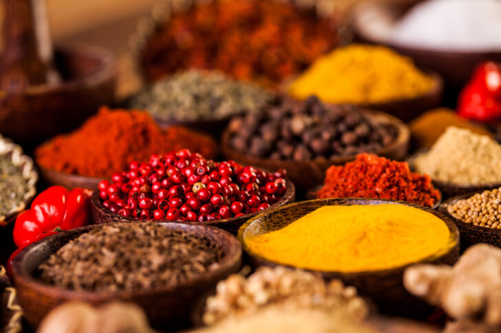 Assorted spices in wooden bowls, including turmeric, paprika, black peppercorns, and pink peppercorns, showcasing rich colors and textures.