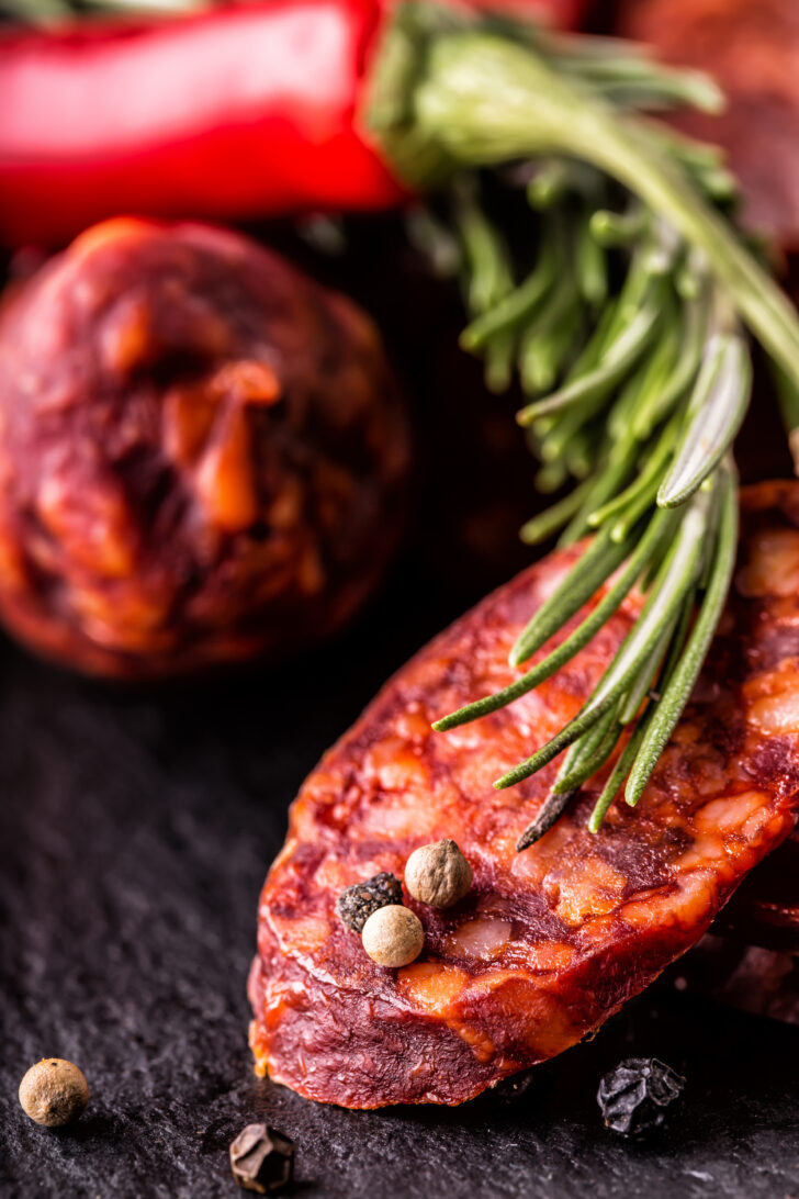 Close-up of sliced chorizo with marbled fat, rosemary, and peppercorns on a dark slate surface, with a red chili pepper in the background.