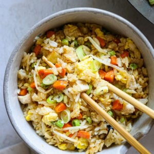 bowl of chicken fried rice with eggs, carrots, peas, sprouts, and scallions, served with chopsticks on a neutral background