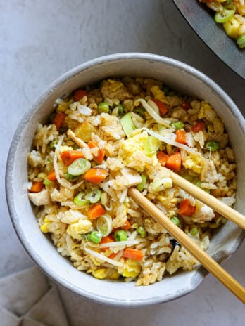 bowl of chicken fried rice with eggs, carrots, peas, sprouts, and scallions, served with chopsticks on a neutral background
