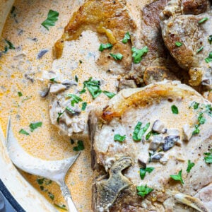Seared pork chops simmering in a creamy mushroom sauce, garnished with chopped parsley and ready to serve in a white pan