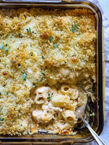 Golden baked mac and cheese in a glass dish with a crispy breadcrumb topping, freshly garnished with herbs and a spoonful served from the corner.