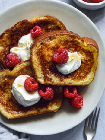 Golden French toast on a plate, topped with whipped cream, raspberries, and maple syrup, ready to serve.