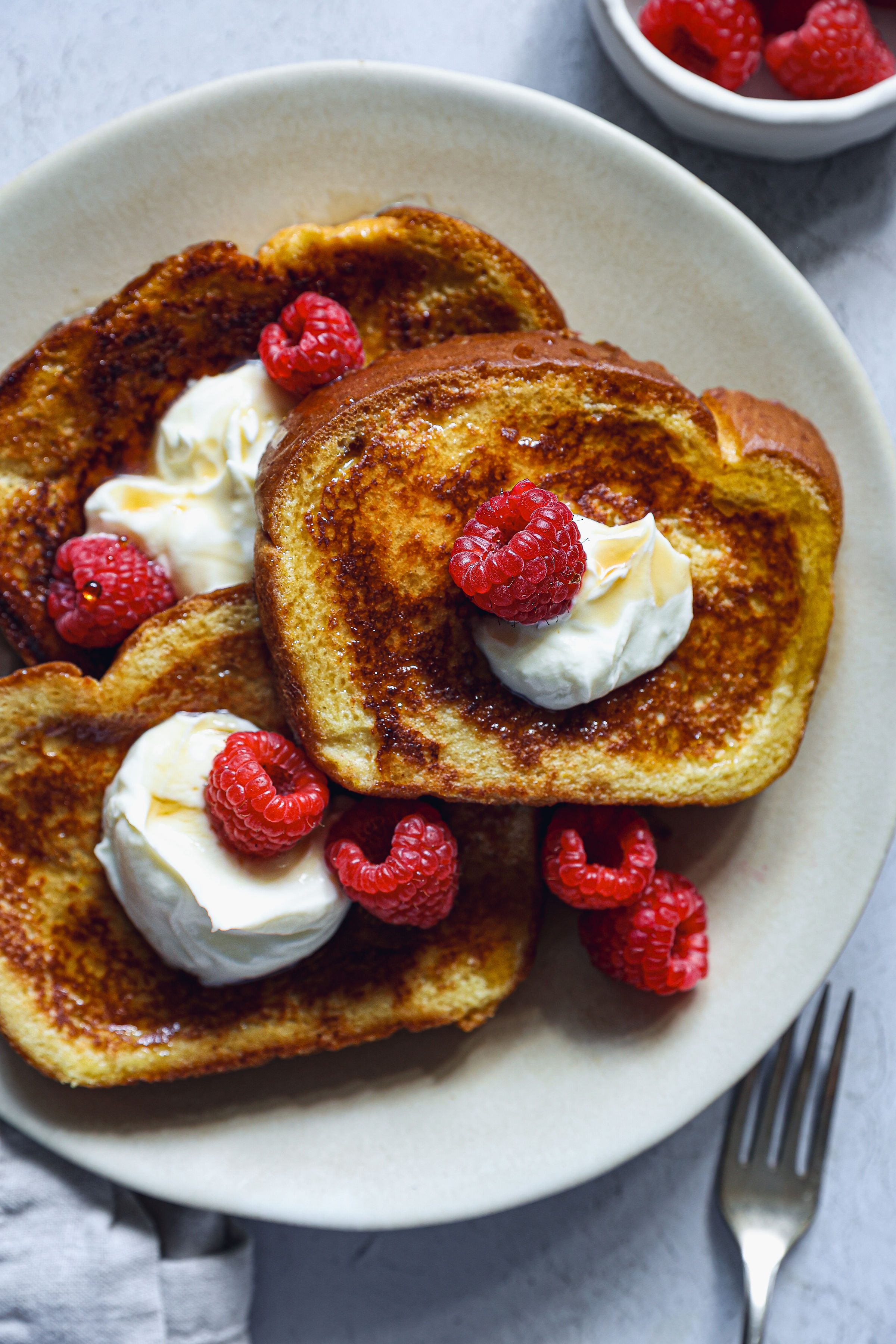Golden French toast on a plate, topped with whipped cream, raspberries, and maple syrup, ready to se