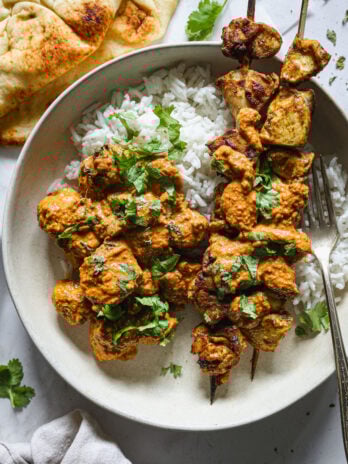 Butter chicken with sauce over rice and grilled skewers, garnished with cilantro and served with naan in a shallow ceramic bowl.