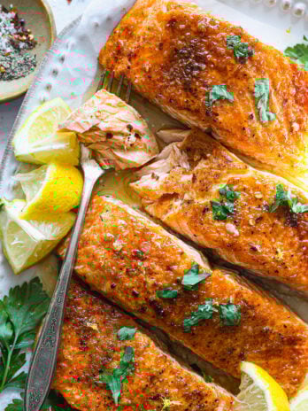 Fork-tender salmon flaked open to reveal juicy, glazed flesh, with lemon wedges and fresh parsley on a white serving plate.