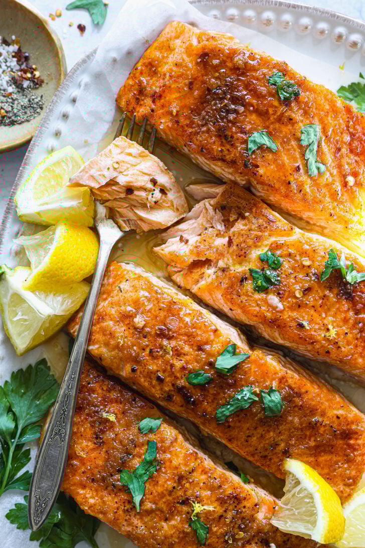Fork-tender salmon flaked open to reveal juicy, glazed flesh, with lemon wedges and fresh parsley on a white serving plate.
