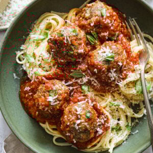 Plated spaghetti topped with tomato sauce, meatballs, fresh basil, and grated Parmesan, served in a green bowl with a fork.