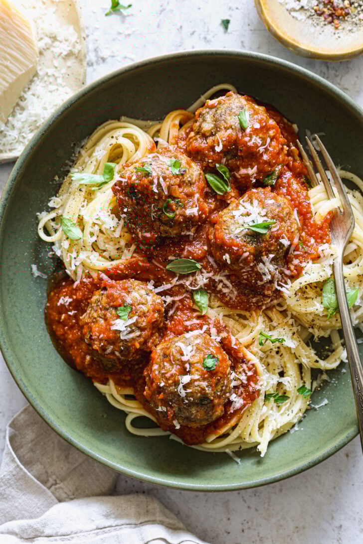 Plated spaghetti topped with tomato sauce, meatballs, fresh basil, and grated Parmesan, served in a green bowl with a fork.