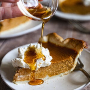 Pumpkin pie topped with maple whipped cream and spices, with one slice cut and lifted from the dish on a pie server.