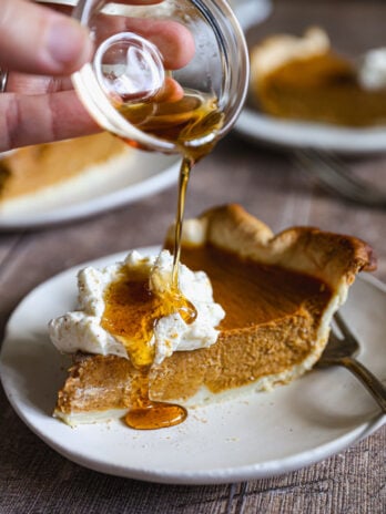 Pumpkin pie topped with maple whipped cream and spices, with one slice cut and lifted from the dish on a pie server.