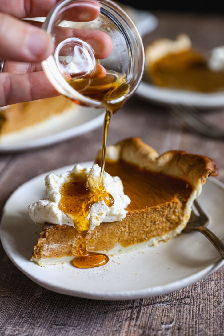 Pumpkin pie topped with maple whipped cream and spices, with one slice cut and lifted from the dish on a pie server.
