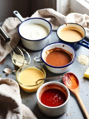 Top-down view of five French mother sauces in rustic pots on a messy kitchen counter with utensils, flour, and herbs scattered around.