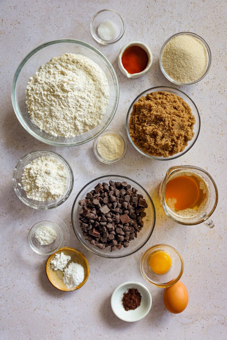 Overhead view of cookie ingredients: flours, sugars, brown butter, eggs, chocolate chips, vanilla, dry milk, and cornstarch