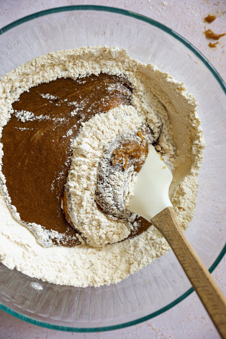 Folding melted butter mixture into flour with a spatula to form chocolate chip cookie dough in a large glass mixing bowl