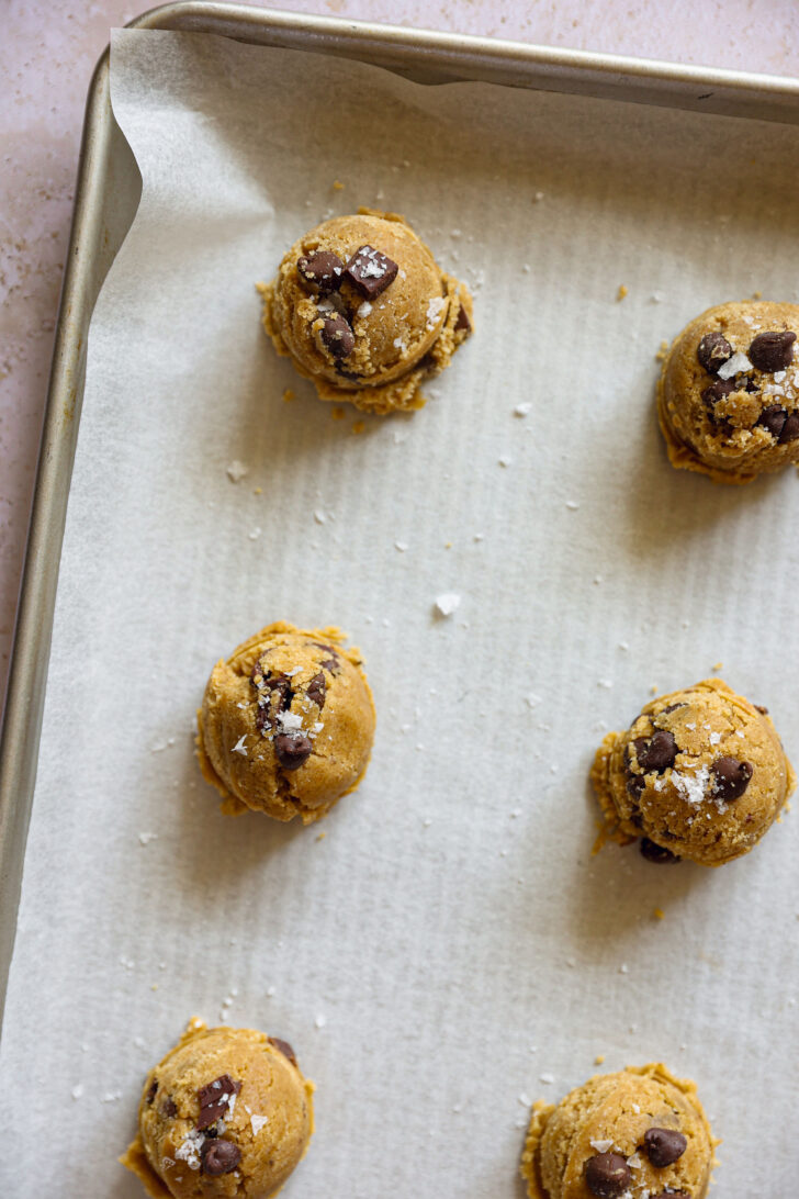 Scooped cookie dough on a parchment-lined baking sheet sprinkled with flaky salt, ready to bake into chocolate chip cookies