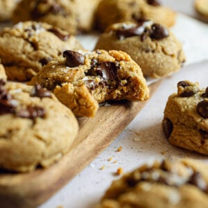Close-up of soft chocolate chip cookies on a wood board, with one broken open to show the gooey, melty center and flaky salt finish