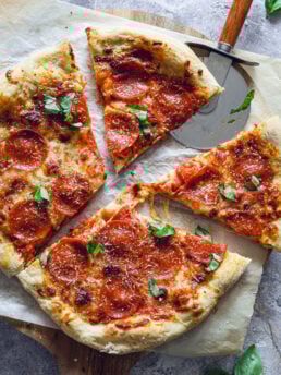 Sliced pepperoni pizza with melted cheese and basil on parchment, shown with a pizza cutter on a wooden peel.