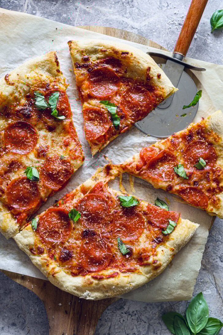 Sliced pepperoni pizza with melted cheese and basil on parchment, shown with a pizza cutter on a wooden peel.