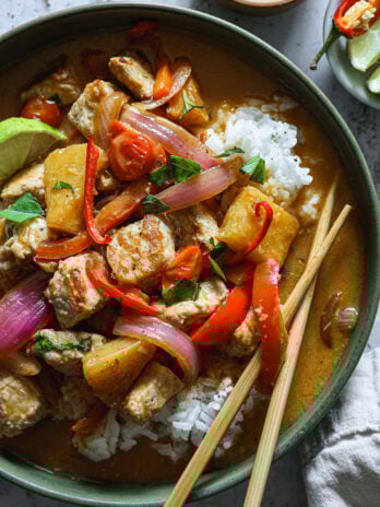 Close-up of Thai red curry pork served over rice, with pineapple, red pepper, onion, tomatoes, and basil, garnished with lime and chili slices.